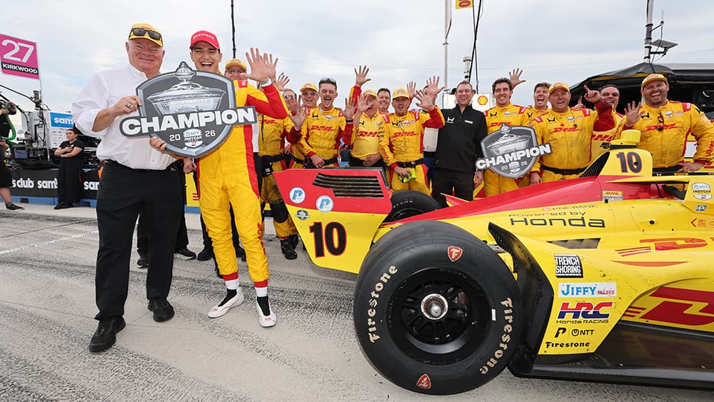Chip Ganassi and Alex Palou celebrating with a 2026 champion sign with Palou holding up five fingers signaling his fifth championship.