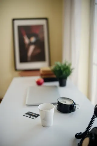 Desk workspace featuring coffee cup, laptop, and plant with artwork in background during a sunny day
